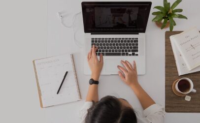 Overhead view of a person typing on a laptop beside a notebook and coffee